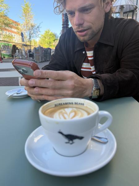 A man is engrossed in his smartphone while enjoying a latte with heart-shaped latte art at an outdoor cafe